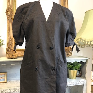 以下が含まれることがあります： 黒いボタンがついたダブルブレストのダークグレーのリネンブレザー。ブレザーは肩と袖口に装飾的な刺繍のデザインが施された短い袖です。