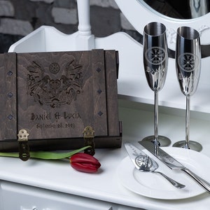 Peut inclure: Une boîte en bois avec un couvercle à charnières, gravée d'un motif de nœuds vikings et des noms "Daniel & Lucia" ainsi que la date "September 28, 2023". Deux flûtes à champagne argentées avec des motifs de boussole viking gravés et un couteau à gâteau argenté avec un motif de boussole viking sont sur une assiette blanche.