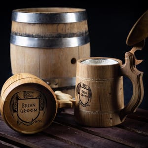 May include: Wooden beer stein with a hinged lid and a decorative handle. The stein has a custom engraved crest with the text "Brian Groom". A small wooden barrel is in the background. The items are made of light brown wood.