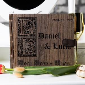 May include: A wooden photo album with a brown leather strap. The album is engraved with the names "Daniel & Lucia" and the date "October 21, 2023". The album also features a decorative "E" and "L" with floral designs and the text "Once Upon a Time..."