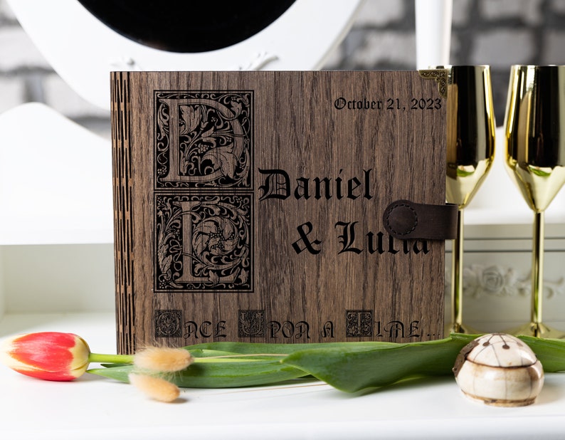 May include: A wooden photo album with a brown leather strap. The album is engraved with the names "Daniel & Lucia" and the date "October 21, 2023". The album also features a decorative "E" and "L" with floral designs and the text "Once Upon a Time..."