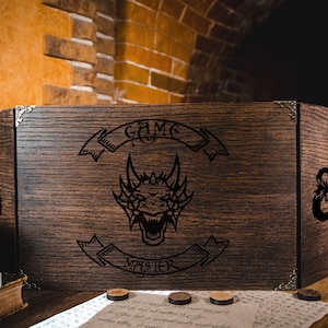 May include: A wooden game master screen with a dragon and the text "Game Master" engraved on the front. The screen has two panels on each side and a D&D symbol on each side panel.