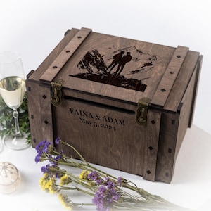 May include: A brown wooden wedding card box featuring an engraved mountain scene and couple silhouette on the lid. The box has metal clasps and the names FAINA & ADAM and the date May 3, 2024 are engraved on the front.