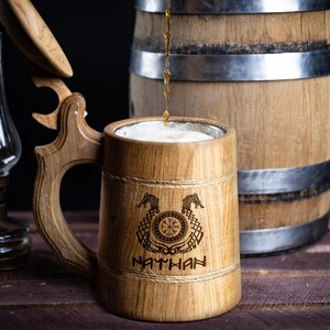 May include: A wooden beer stein with a lid, engraved with a Viking compass and two dragons. The stein is filled with beer and the name "NATHAN" is engraved below the design.