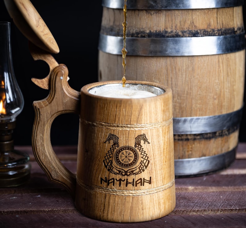 May include: A wooden beer stein with a lid, engraved with a Viking compass and two dragons. The stein is filled with beer and the name "NATHAN" is engraved below the design.