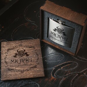 Puede incluir: Una caja de madera con tapa abatible, que contiene una petaca de plata con un dise&ntilde;o grabado de un dado de Dungeons and Dragons y el texto "Michael Dungeon Master". La caja tambi&eacute;n est&aacute; grabada con el mismo dise&ntilde;o y texto.