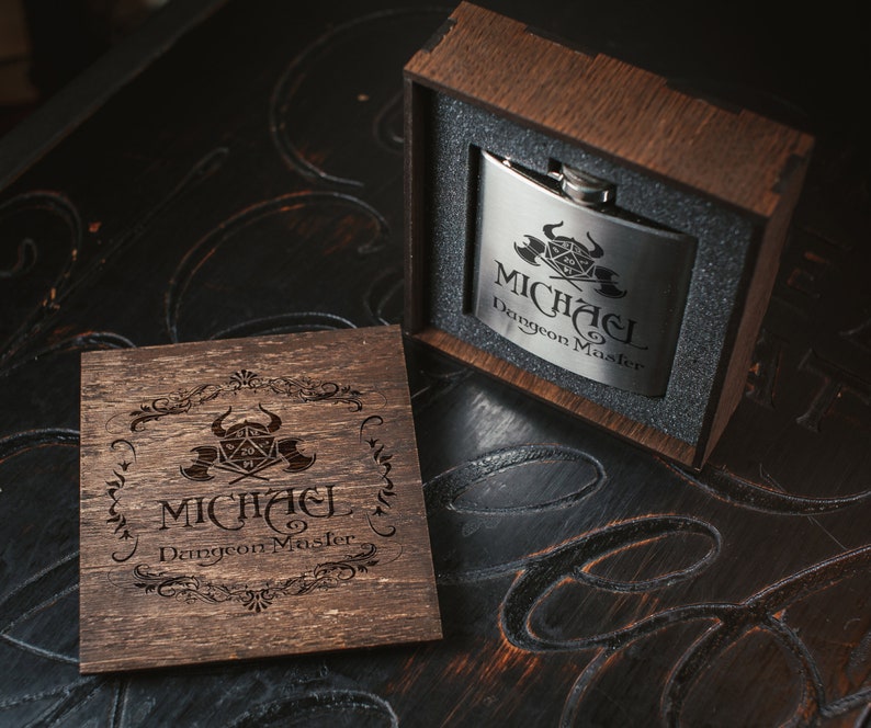 Puede incluir: Una caja de madera con tapa abatible, que contiene una petaca de plata con un dise&ntilde;o grabado de un dado de Dungeons and Dragons y el texto "Michael Dungeon Master". La caja tambi&eacute;n est&aacute; grabada con el mismo dise&ntilde;o y texto.