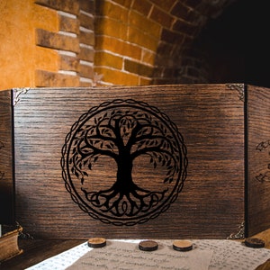 Könnte beinhalten: Eine Holzkiste mit einem Scharnierdeckel, verziert mit einem schwarzen und weißen Baum des Lebens. Die Kiste hat Metallverzierungen und ist geschlossen.