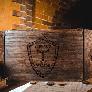 Può includere: Schermo da gioco da tavolo in legno scuro con la scritta incisa "CHOOSE YOUR WEAPON". Lo schermo presenta dettagli angolari in metallo e un disegno a scudo con un simbolo di caduceo. Il legno ha una ricca finitura marrone scuro.
