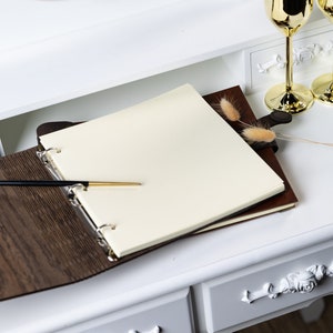 May include: A brown leather-bound guest book with a blank white page open, a gold pen lying on the page, and a few dried flowers on the cover.