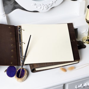 May include: A brown leather-bound guest book with a blank white page open, a black pen, and a small blue velvet box with a gold lid.