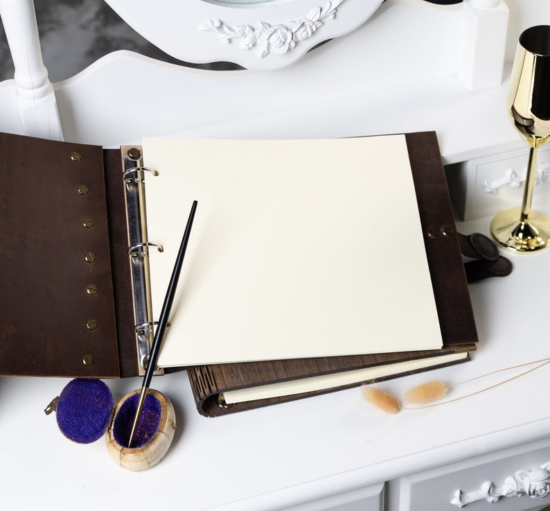 May include: A brown leather-bound guest book with a blank white page open, a black pen, and a small blue velvet box with a gold lid.