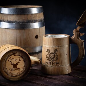 May include: Two wooden beer steins with lids. One stein has a deer and the text "Eric Groom" engraved on it. The other stein has a Viking knot design and the text "Nathan" engraved on it. Both steins are sitting on a wooden surface next to a wooden barrel.