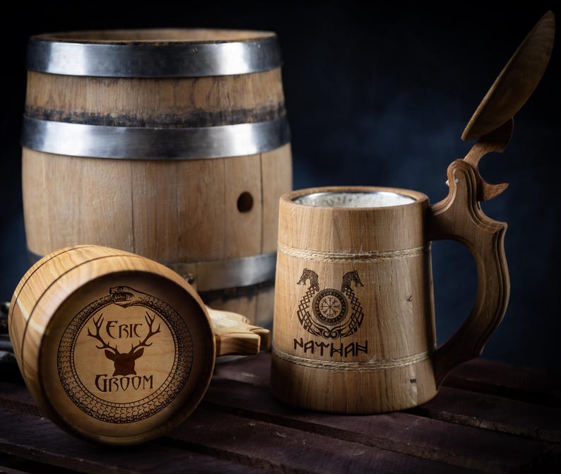 May include: Two wooden beer steins with lids. One stein has a deer and the text "Eric Groom" engraved on it. The other stein has a Viking knot design and the text "Nathan" engraved on it. Both steins are sitting on a wooden surface next to a wooden barrel.