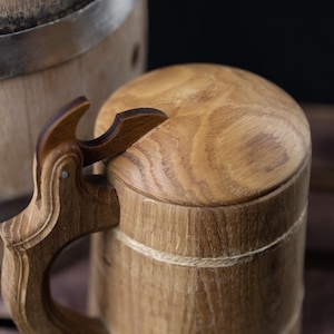 May include: A wooden beer stein with a hinged lid and a carved handle. The stein is made of light brown wood and has a natural finish. The lid is secured with a rope.