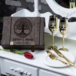 Könnte beinhalten: Eine Holzkiste mit einem eingravierten Lebensbaum auf dem Deckel. Die Kiste ist geschlossen und hat zwei Messingverschlüsse. Die Kiste ist mit "Kevin & Anna, 12. Dezember 2021" beschriftet. Zwei goldene Champagnerflöten mit einem Lebensbaumdesign stehen neben der Kiste. Ein goldenes Kuchmesser und eine Serviergabel liegen auf einem weißen Teller.