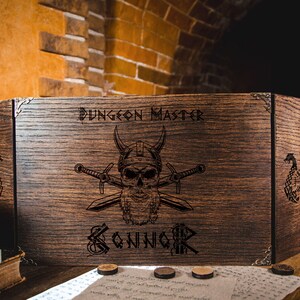 May include: A wooden Dungeon Master screen featuring a skull and crossed swords design. The screen is engraved with the words "Dungeon Master" and the name "Konnor". The screen is brown with metal corner accents.