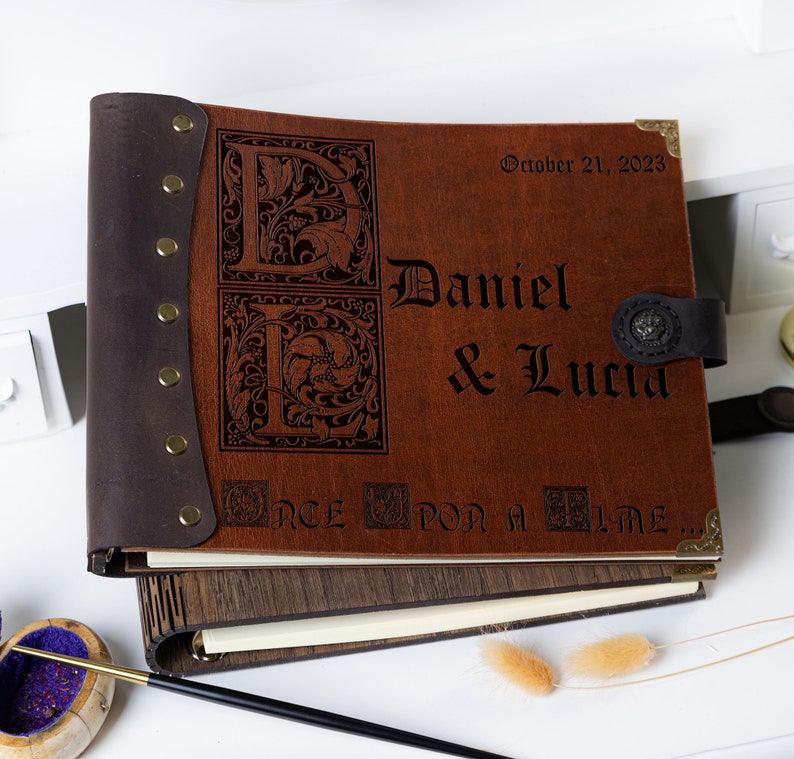 May include: A brown leather-bound photo album with a wooden cover. The album is decorated with a metal clasp and a metal plate with the text "Daniel & Lucia" and the date "October 21, 2023". The album also has the text "Once Upon a Time..." engraved on the front.