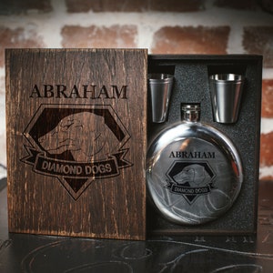 Könnte beinhalten: Eine Holzkiste mit einem Scharnierdeckel, die eine Edelstahl-Trinkflasche mit der Gravur "Abraham Diamond Dogs" enthält. Die Flasche wird von zwei kleinen Schnapsgläsern begleitet.