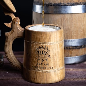 May include: A wooden beer stein with a dragon eye design and the text "You can certainly try". The stein has a hinged lid and a handle. It is being filled with a dark liquid from a wooden barrel.