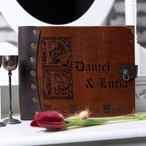 May include: A brown leather-bound photo album with a personalized inscription. The album features a decorative design with the initials "D" and "L" and the date "October 21, 2023". The inscription reads "Daniel & Lucia".