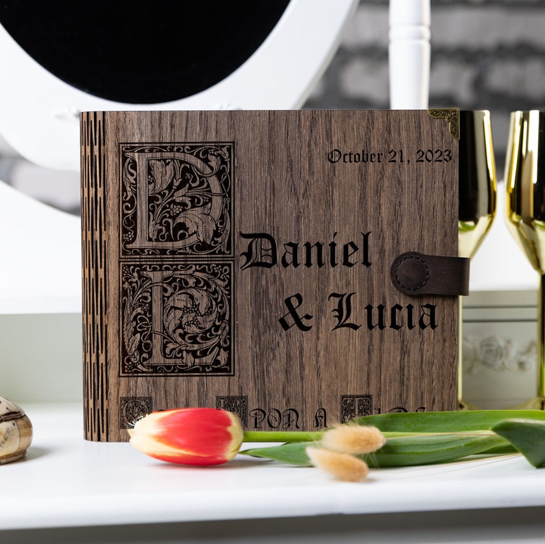 May include: A wooden photo album with a dark brown finish. The album is engraved with the names "Daniel & Lucia" and the date "October 21, 2023". The initials "D & L" are also engraved in a decorative font.