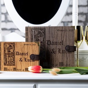 May include: Two wooden photo albums with engraved initials and the names "Daniel & Lucia" and the date "October 21, 2023". The albums are closed and have a leather strap.