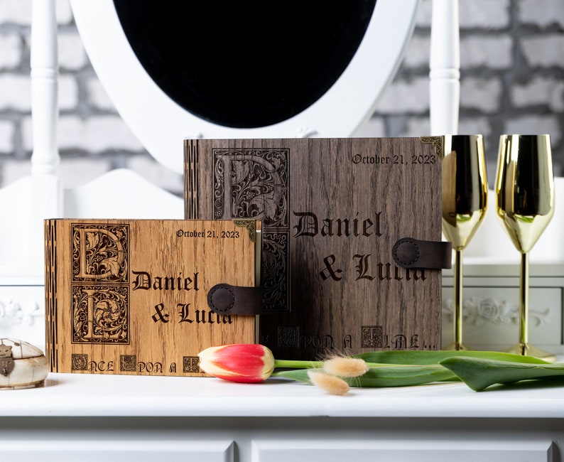 May include: Two wooden photo albums with engraved initials and the names "Daniel & Lucia" and the date "October 21, 2023". The albums are closed and have a leather strap.