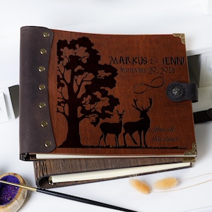 May include: A brown leather photo album with a wood cover. The front cover features an engraved design of a tree, two deer, and the text "Markus & Jenni September 20, 2025 After all this time?"