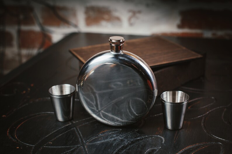 Puede incluir: Una petaca de plata con un cuerpo redondo y plano y una tapa de plata. Dos peque&ntilde;os vasos de chupito de plata est&aacute;n a cada lado de la petaca.