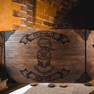 Op de afbeelding: Een bruin houten Dungeon Master-scherm met gegraveerde ontwerpen. Het scherm toont een ridderhelm, de woorden "Dungeon Master" en Keltische knoophoeken. Het scherm staat op een houten tafel.