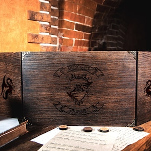 May include: A wooden Dungeon Master screen with the words "Dungeon Master" and a crest etched into the front. The screen is dark brown with metal corner protectors and dragon silhouettes. A mortar and pestle, dice, and a scroll are on the table.