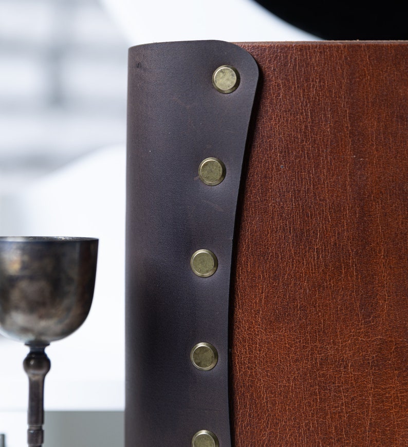 May include: A brown leather-bound journal with a dark brown leather flap and decorative brass rivets.