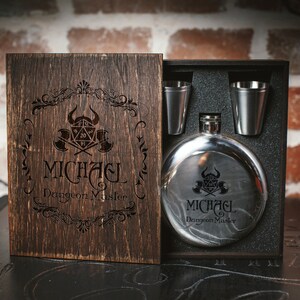 Puede incluir: Una caja de madera con tapa abatible, que contiene una petaca de plata y dos vasos de chupito. La petaca y los vasos est&aacute;n grabados con un dise&ntilde;o de tem&aacute;tica de Dungeons and Dragons y el nombre "MICHAEL Dungeon Master".