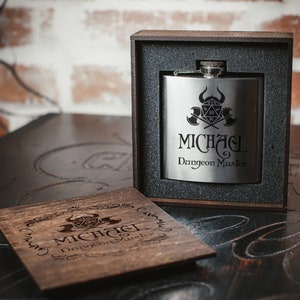 Puede incluir: Una petaca de plata con un dise&ntilde;o grabado en negro de un dado, hachas y el texto "MICHAEL Dungeon Master". La petaca est&aacute; en una caja de madera con un dise&ntilde;o grabado similar.