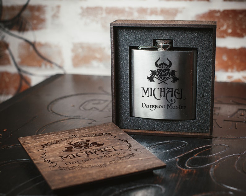 Puede incluir: Una petaca de plata con un dise&ntilde;o grabado en negro de un dado, hachas y el texto "MICHAEL Dungeon Master". La petaca est&aacute; en una caja de madera con un dise&ntilde;o grabado similar.