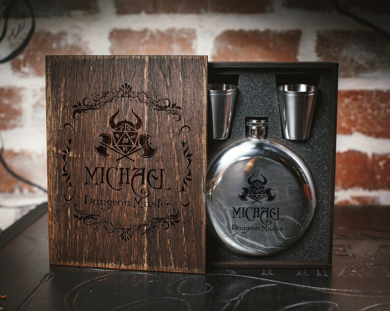 Puede incluir: Una caja de madera con tapa con bisagras, que contiene una petaca de plata con un dise&ntilde;o en blanco y negro y dos vasos de chupito de plata. La petaca y los vasos de chupito est&aacute;n grabados con el texto "MICHAEL Dungeon Master" y un s&iacute;mbolo de Dungeons and Dragons.