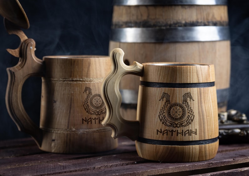 May include: Two wooden beer steins with black bands and engraved designs. The steins have the names "Nathan" and "NATH" engraved on them. The steins are sitting on a wooden surface.