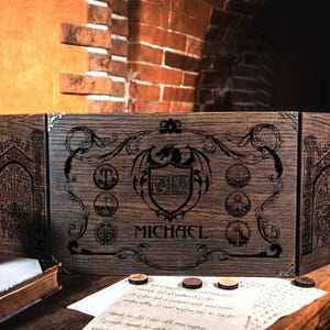Peut inclure: Une boîte pliante en bois à trois panneaux avec un blason de dragon et le nom "MICHAEL" gravé sur le devant. La boîte est en bois foncé et présente des sculptures complexes.