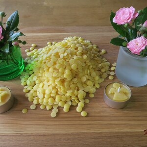 Pu&ograve; includere: Un mucchio di granuli di cera d'api gialla su una superficie di legno con tre piccole candele, una a forma di cuore, una a forma di stella e una a forma di angelo. Due piccoli vasi con rose rosa sono su entrambi i lati della cera d'api.