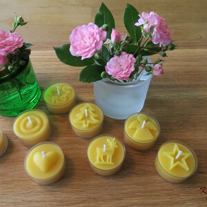 May include: Assortment of eight yellow beeswax tea light candles with various designs, including a snowflake, a heart, a Christmas tree, a deer, an angel, a star, and a circle. The candles are arranged on a wooden surface with two small vases of pink roses.