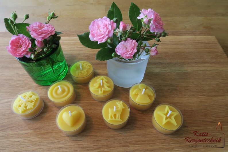 May include: Assortment of eight yellow beeswax tea light candles with various designs, including a snowflake, a heart, a Christmas tree, a deer, an angel, a star, and a circle. The candles are arranged on a wooden surface with two small vases of pink roses.