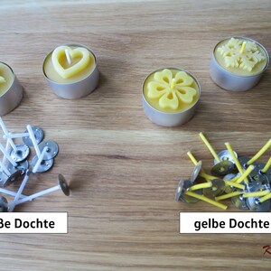 May include: Four tealight candles with different designs, including a star, heart, flower, and snowflake.  The candles are made of beeswax and are surrounded by white and yellow wicks with metal bases. The text "wei&szlig;e Dochte" and "gelbe Dochte" is visible below the wicks.