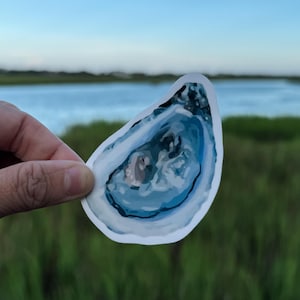 May include: A sticker featuring a stylized oyster shell design. The shell is primarily shades of blue and white, with a detailed interior. The sticker is held against a backdrop of a body of water and green marsh grass.