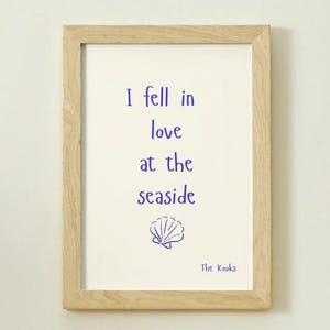 May include: A light wood frame containing a white print with the purple text I fell in love at the seaside, followed by a purple line-art illustration of a seashell and the text The Kooks in the bottom right corner of the print.