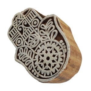 Könnte beinhalten: Ein Holzblock-Stempel mit einem detaillierten Design einer Hamsa-Hand, einem Symbol für Schutz und Glück. Das Design zeigt komplizierte florale Muster und geometrische Formen in Weiß auf einem braunen Holzgrund.