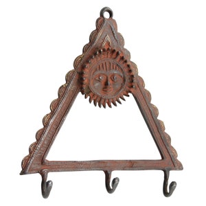 Może przedstawiać: Trójkątny wieszak na klucze z trzema haczykami, wykonany z metalu o rdzawo-brązowym wykończeniu. Górna część wieszaka przedstawia słońce z twarzą, z złotymi akcentami wokół krawędzi.