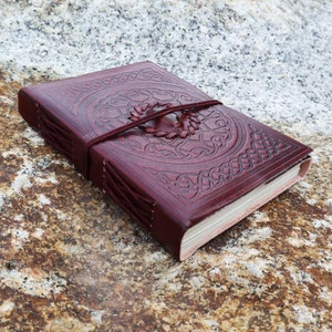 May include: A burgundy leather journal with a Celtic knot design embossed on the cover. The journal is closed and has a leather strap with a metal clasp.