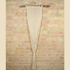 Può includere: Un arazzo da parete in macramè bianco con un bastone di legno in alto e un lungo fiocco in basso. L'arazzo è contro un muro di mattoni.