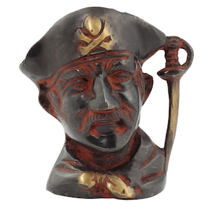 Può includere: Una statuetta di testa di pirata nera e dorata con un teschio e ossa incrociate sul cappello. La statuetta tiene una spada nella mano destra.